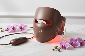 LED Light Therapy Facial Beauty Mask glowing with red LED lights on white marble surface with purple flowers — at-home skincare for clear glowing skin